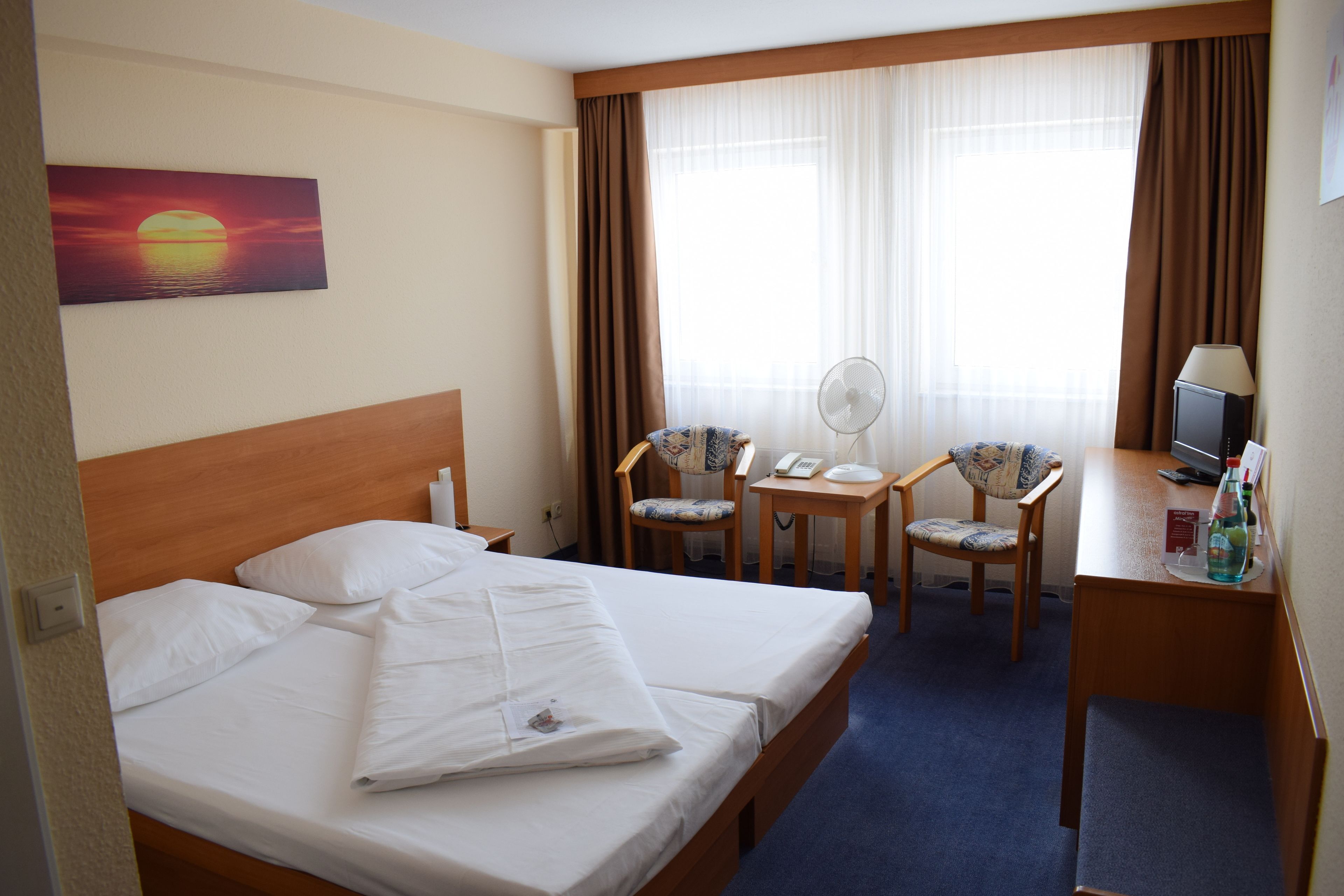 Ein Hotelzimmer mit einem Bett, einem Schreibtisch, zwei Stühlen, einem Fernseher und einem Sonnenuntergangsbild.