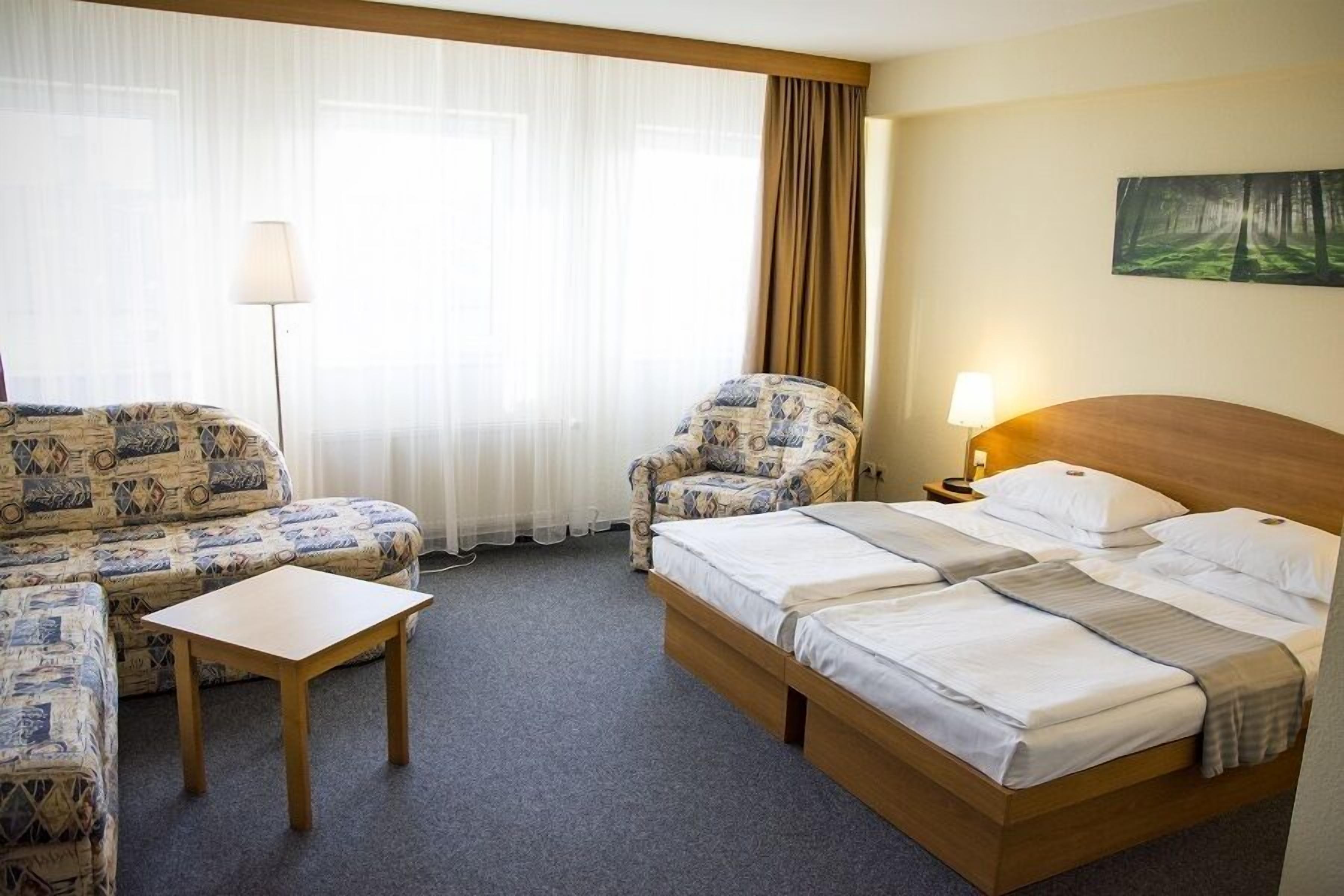 A hotel room with a bed, two patterned sofas, a small table, a lamp, and a painting on the wall.