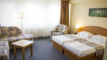 A hotel room with a bed, two patterned sofas, a small table, a lamp, and a painting on the wall.