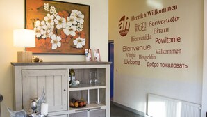 A reception area with a wall-mounted welcome sign in multiple languages, a decorative cabinet with a candle and fruit, and a lamp.