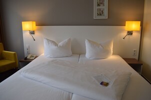 Una cama bien hecha con sábanas blancas, dos lámparas de noche, un teléfono en la mesita y una pequeña mesa con una silla.