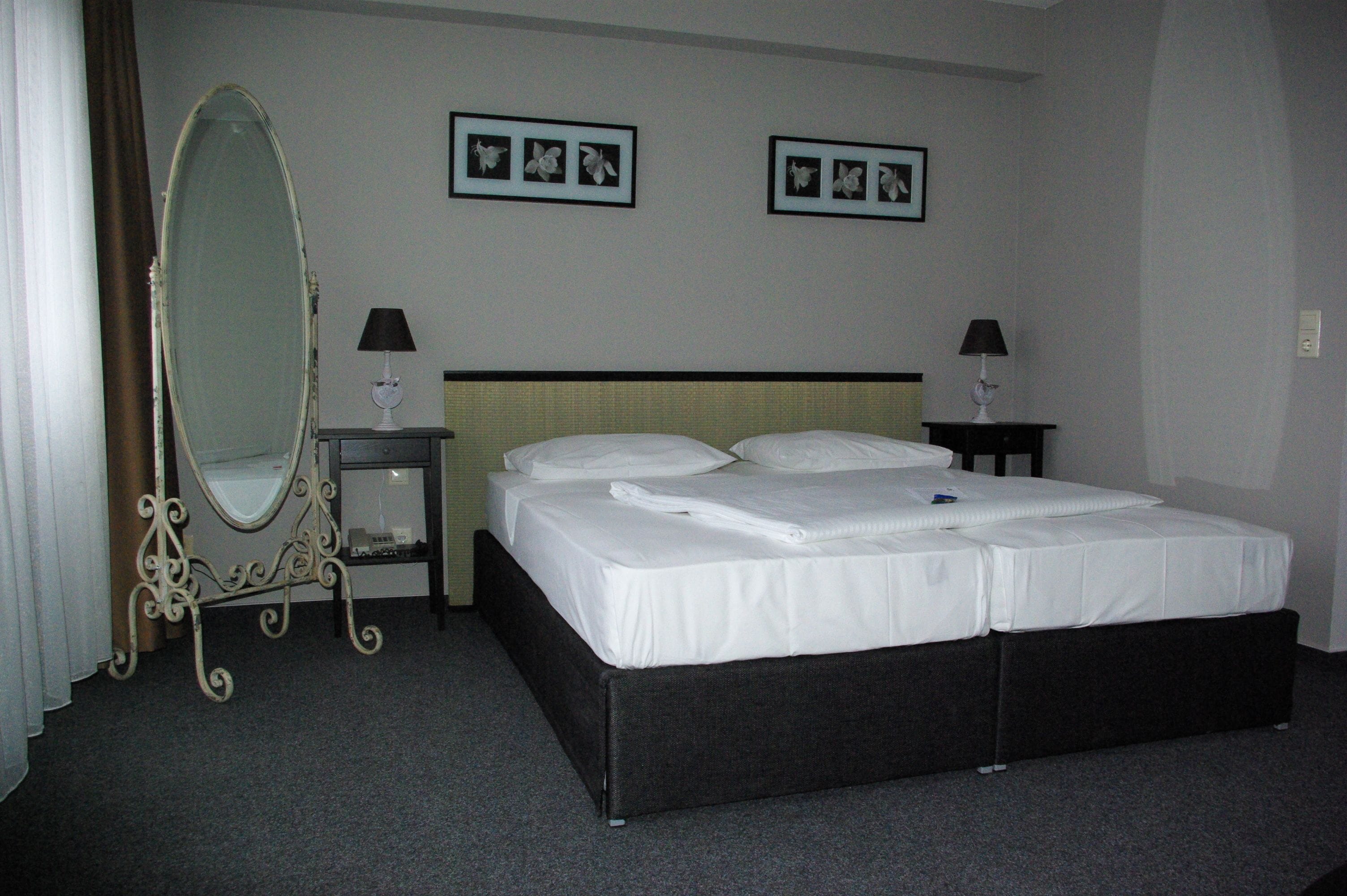 Habitación de hotel con una cama grande, dos mesitas de noche con lámparas, un espejo de cuerpo entero y dos cuadros enmarcados en la pared.