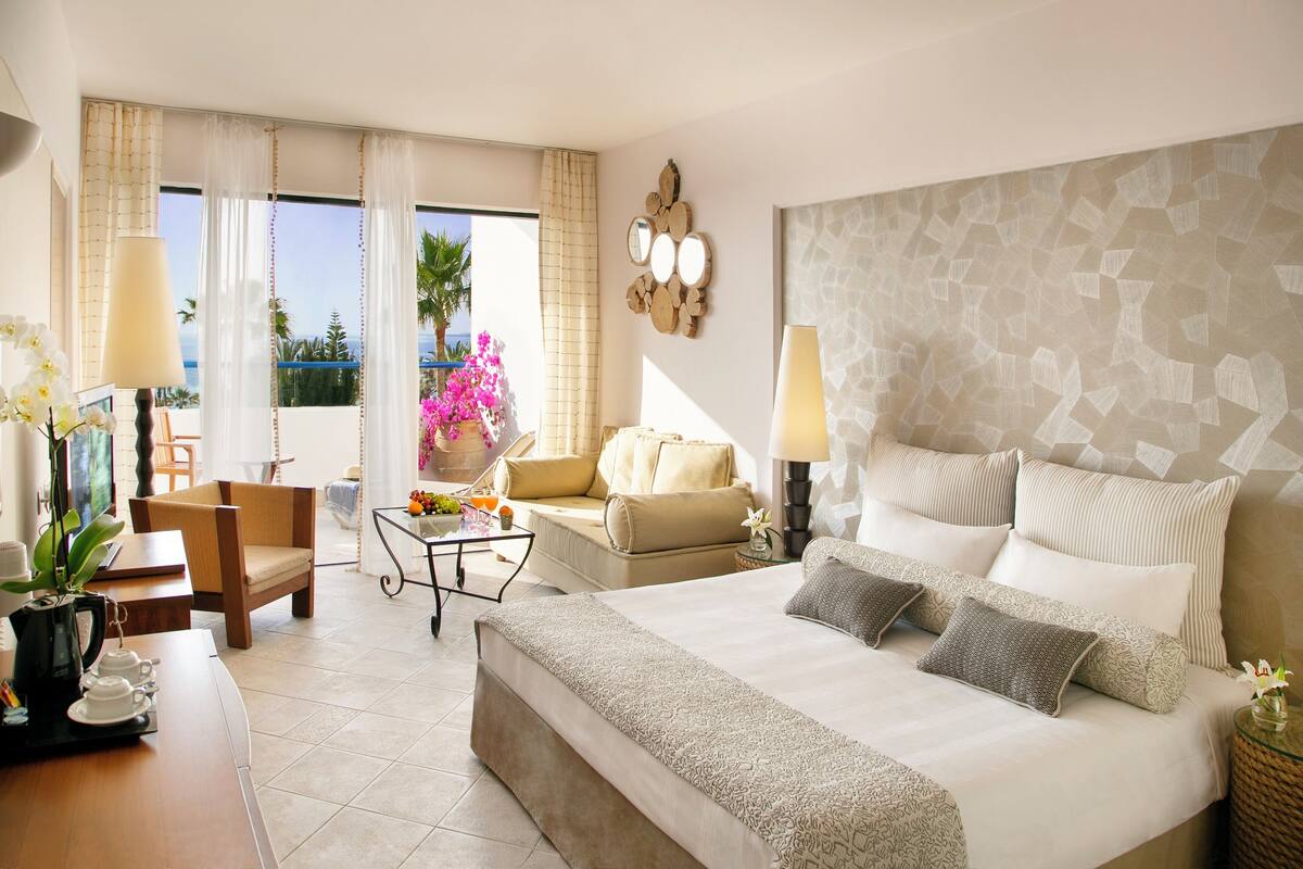 Habitación de hotel con cama, sofá, una mesa de centro y una silla. Tiene una ventana con cortinas, una lámpara y un jarrón con flores.