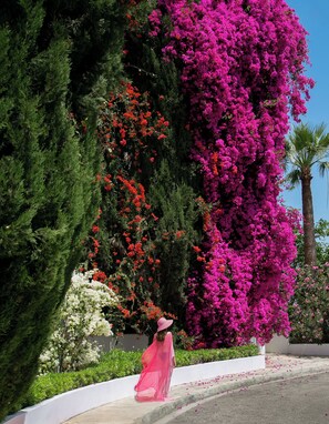 A woman in a pink dress walking along a sidewalk with a garden featuring green trees and pink flowers. - Azia Resort and Spa (Chlorakas)