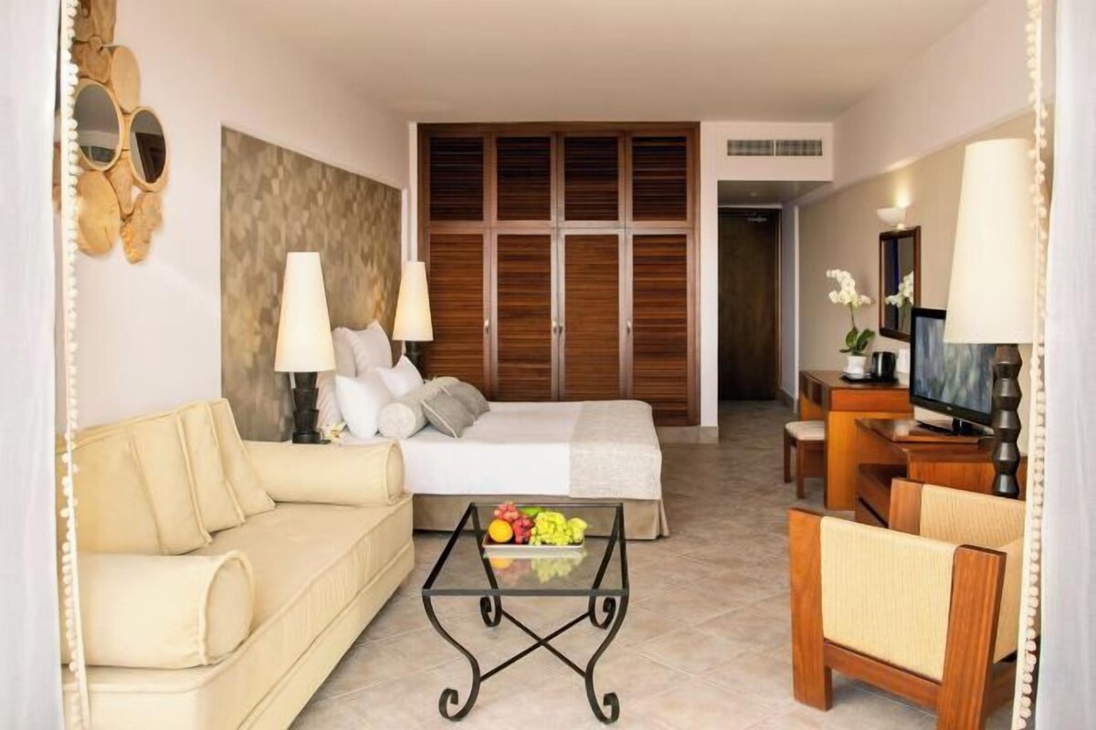 Habitación de hotel con cama, sofá, mesa de centro con fruta, televisión y escritorio con lámpara.