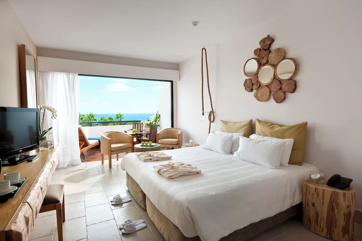 Habitación de hotel con una cama grande, televisión, escritorio con computadora, silla y balcón con vista al océano.