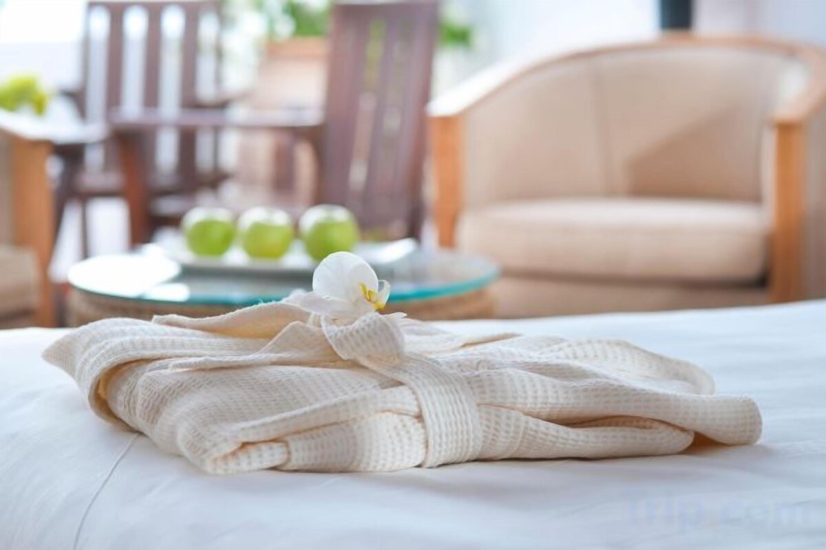 A neatly made bed with a folded towel and a white flower. In the background, there is a round table with green apples and a chair.