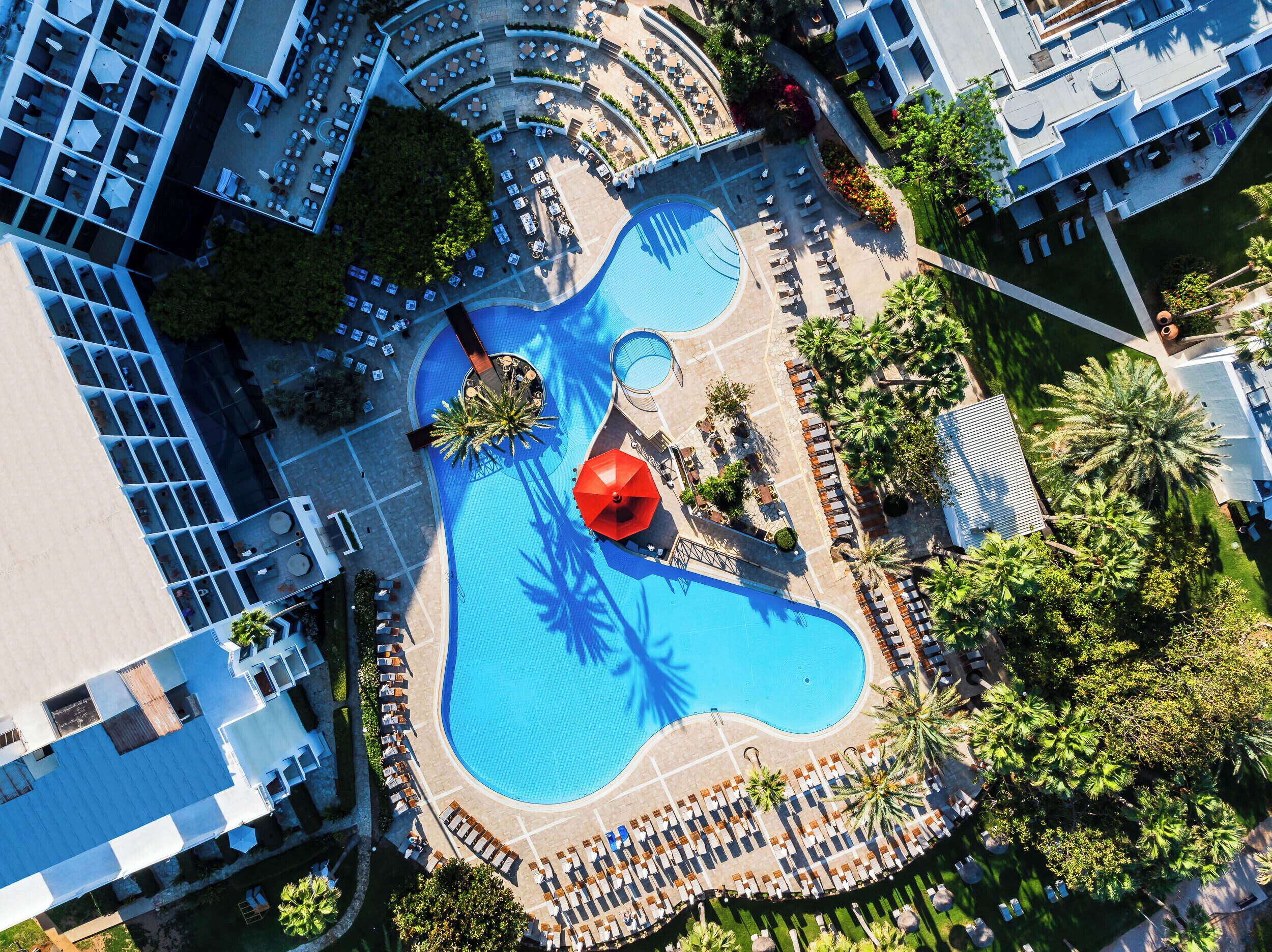 An aerial view of a resort with a central swimming pool, sun loungers, and palm trees.