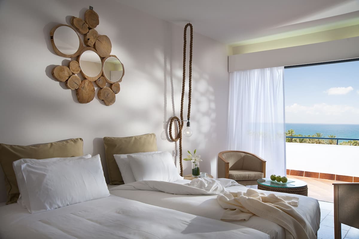 Habitación de hotel con una cama, una silla, una mesita con fruta, un espejo en la pared y vista al océano.
