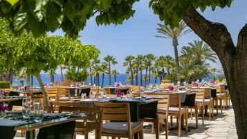 An outdoor restaurant with tables and chairs, palm trees, and a view of the ocean.