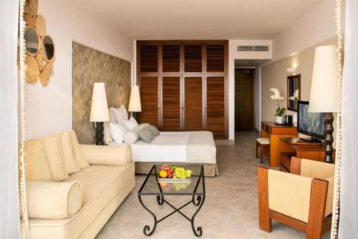 Habitación de hotel con cama, sofá, mesa de centro con fruta, televisor y escritorio.
