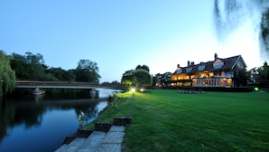 A riverside hotel with a bridge, a well-maintained lawn, and a large building with multiple windows and a chimney.