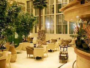 Un vaste espace intérieur aménagé avec différents types de sièges, notamment des chaises et des tables, et agrémenté de nombreuses plantes en pot ainsi que d’une fontaine décorative centrale.