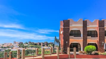 Un edificio histórico con ventanas de arco y fachada roja, con vistas a un paisaje urbano que incluye una colina e iglesia al fondo.