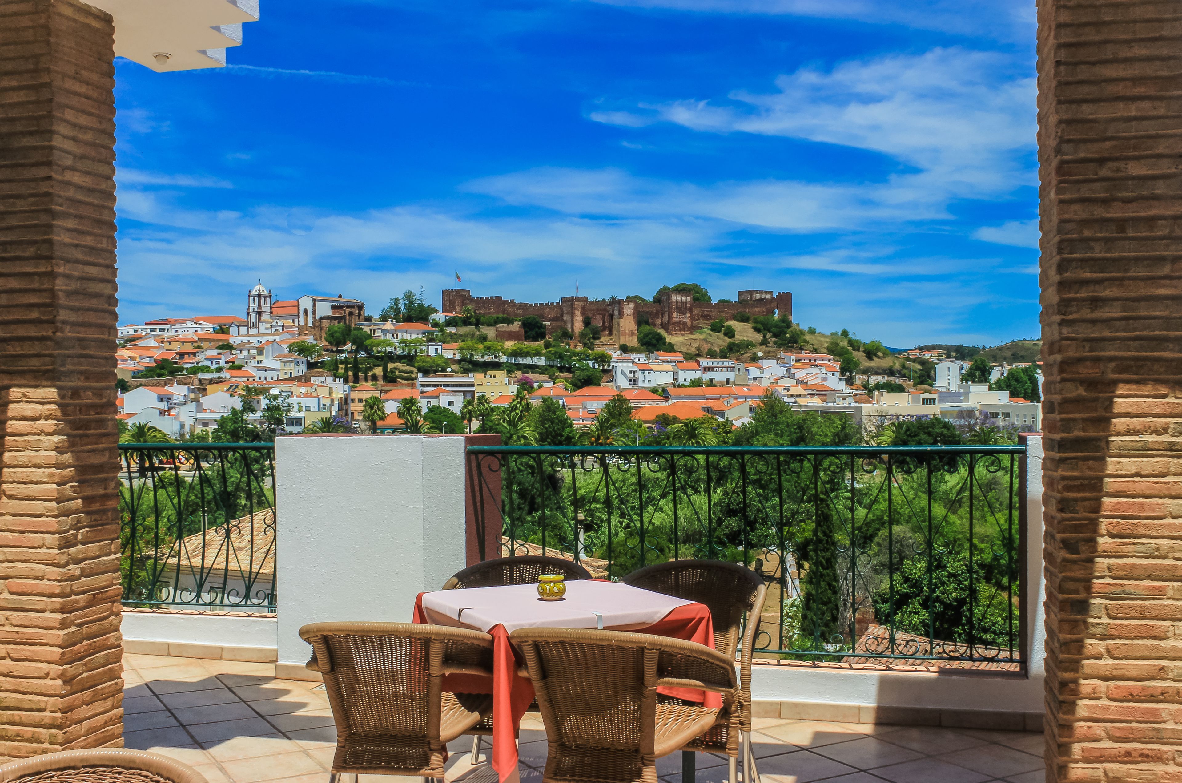A balcony with wicker chairs and a table, overlooking a historic town with a castle and red-roofed buildings.