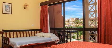 A room with two beds, a window with a view of buildings, a wall-mounted lamp, and a framed picture.