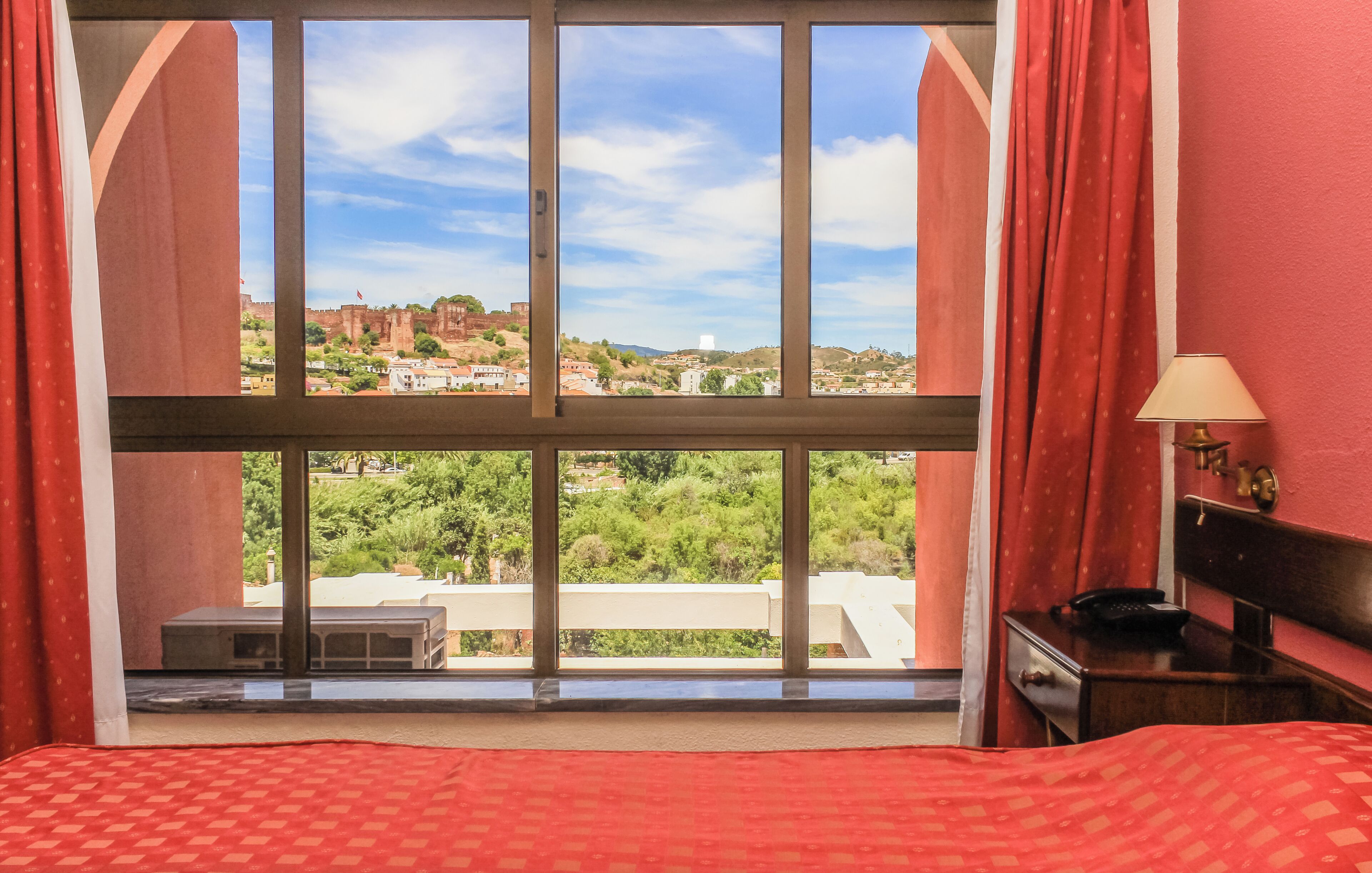 A hotel room with a large window, a bed, a bedside table, and a lamp.