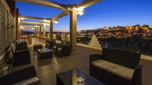 En udendørs terrasse med siddepladser og udsigt over en historisk by ved skumring.