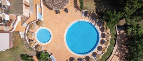 An aerial view of a hotel with two swimming pools, sun loungers, and a covered seating area.
