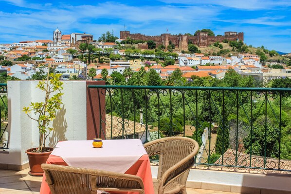 A balcony with wicker chairs and a table, overlooking a town with historic buildings and a castle. - Hotel Colina Dos Mouros (Silves)