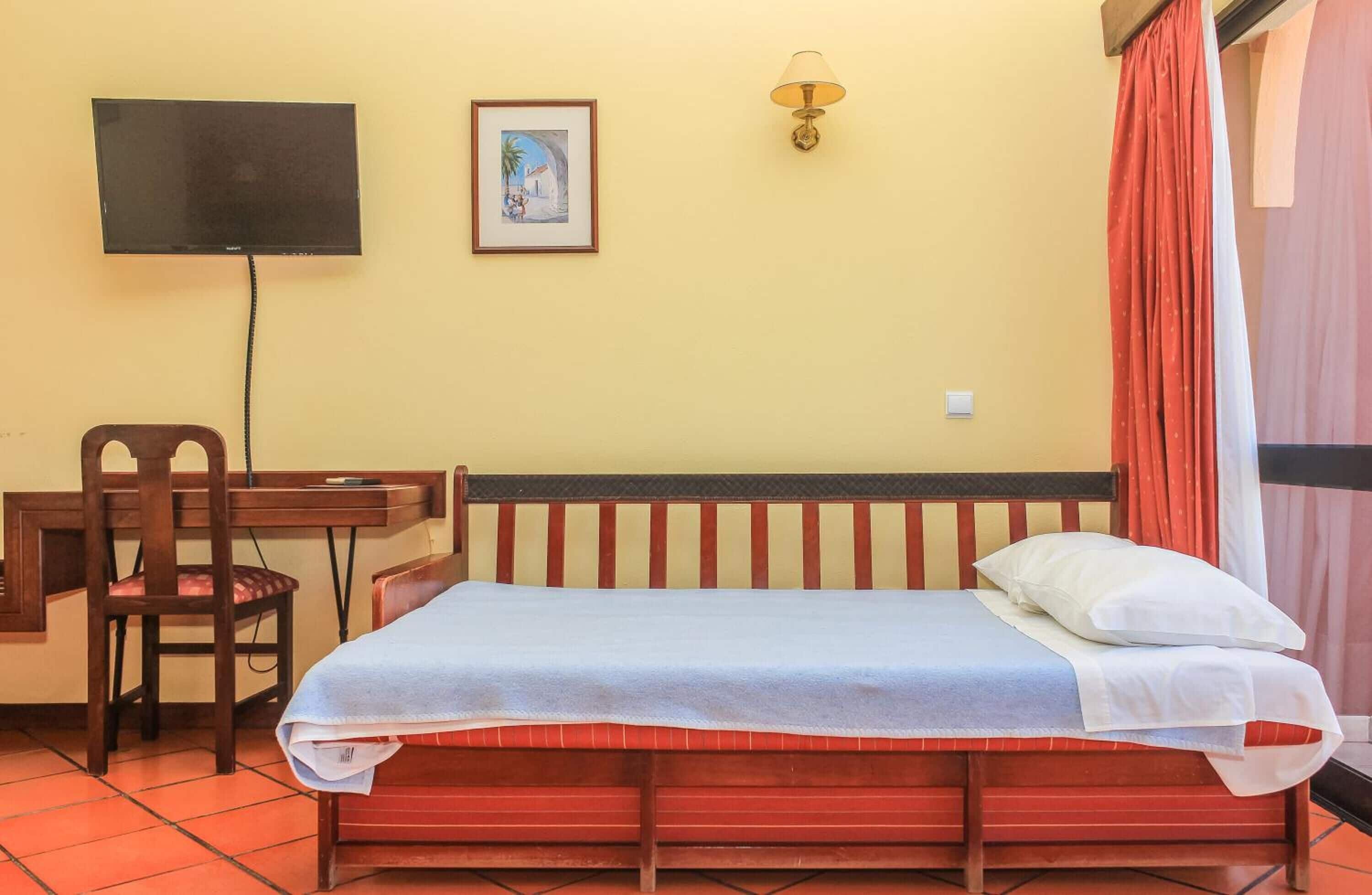 Une pièce comprenant un lit, un bureau, une chaise, un téléviseur et une lampe fixée au mur.