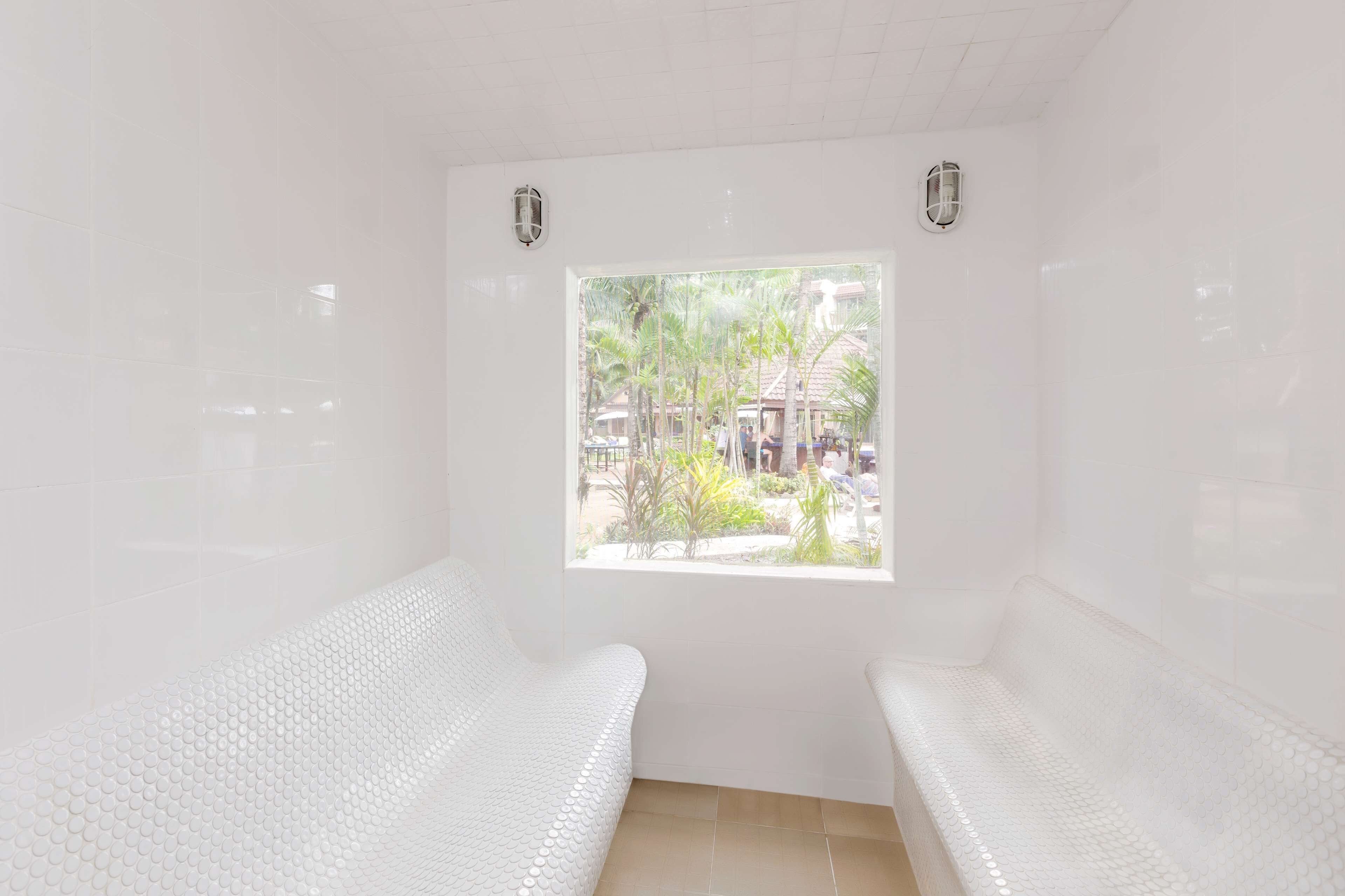 Un cuarto de spa con dos bancos blancos, una ventana que ofrece vistas a palmeras y edificios, y un piso de baldosas.
