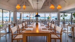 Un comedor amplio con mesas y sillas de madera, ventanas grandes con vista al océano y lámparas colgantes.