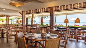 Un restaurante con mesas y sillas de madera, ventanas amplias con vistas al mar, y lámparas decorativas en el techo.