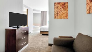 Habitación de hotel con televisión de pantalla plana sobre un tocador de madera, un sofá, una cama con cabecera blanca y dos cuadros abstractos en las paredes.