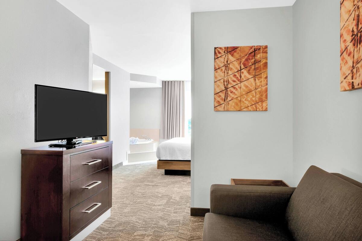 Habitación de hotel con televisión de pantalla plana sobre un tocador de madera, un sofá, una cama con cabecera blanca y dos cuadros abstractos en las paredes.