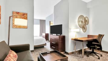 Quarto de hotel com cama, escrivaninha com cadeira, televisão, luminária e um objeto decorativo na parede.