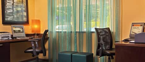 Habitación de hotel con escritorio, dos sillas de oficina, una computadora portátil, una impresora y un pequeño taburete verde.