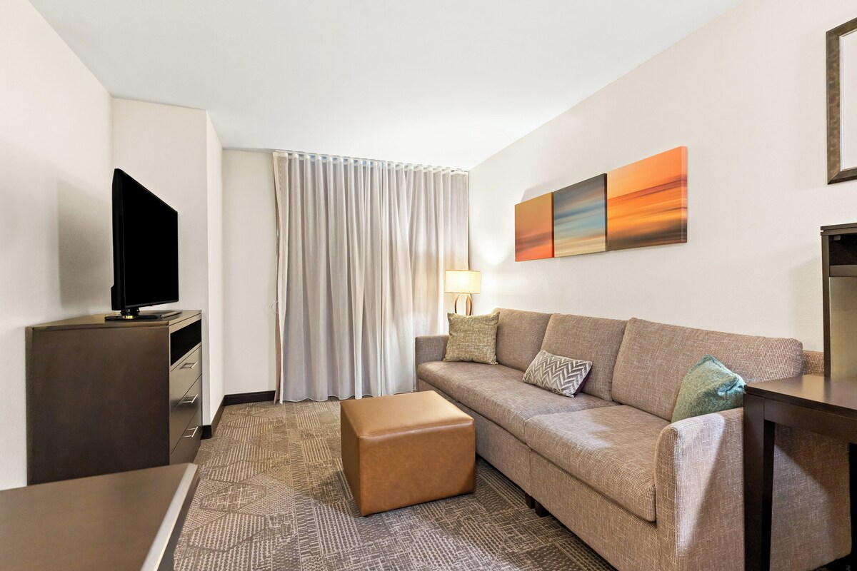 A hotel room with a sofa, ottoman, TV, and artwork on the wall.