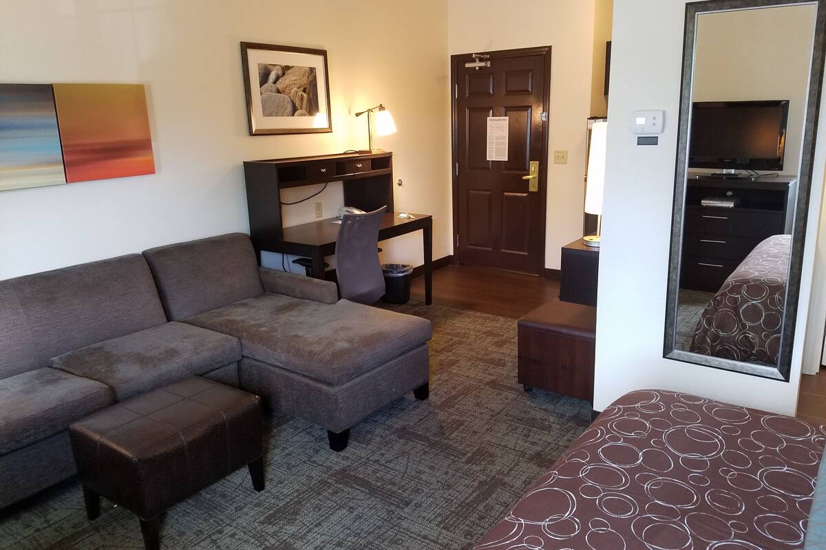 Une chambre d’hôtel comprenant un canapé d’angle, un bureau avec une chaise, une petite table, une poubelle, un lit avec une couvre-lit à motifs, un grand miroir et une porte avec une pancarte.
