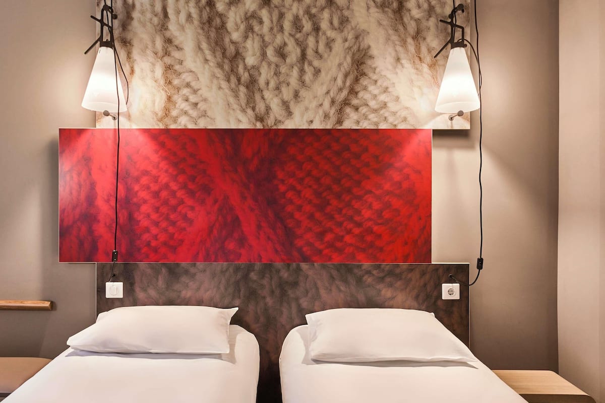 A hotel room with two beds, a large red and brown patterned headboard, and two wall-mounted lamps.