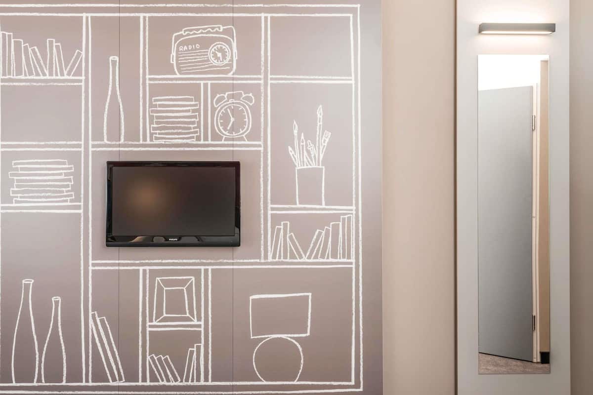 Un mur sur lequel est fixé un téléviseur à écran plat, au-dessus d’une œuvre d’art murale de style tableau noir représentant divers objets, comme des livres, une radio et une horloge.