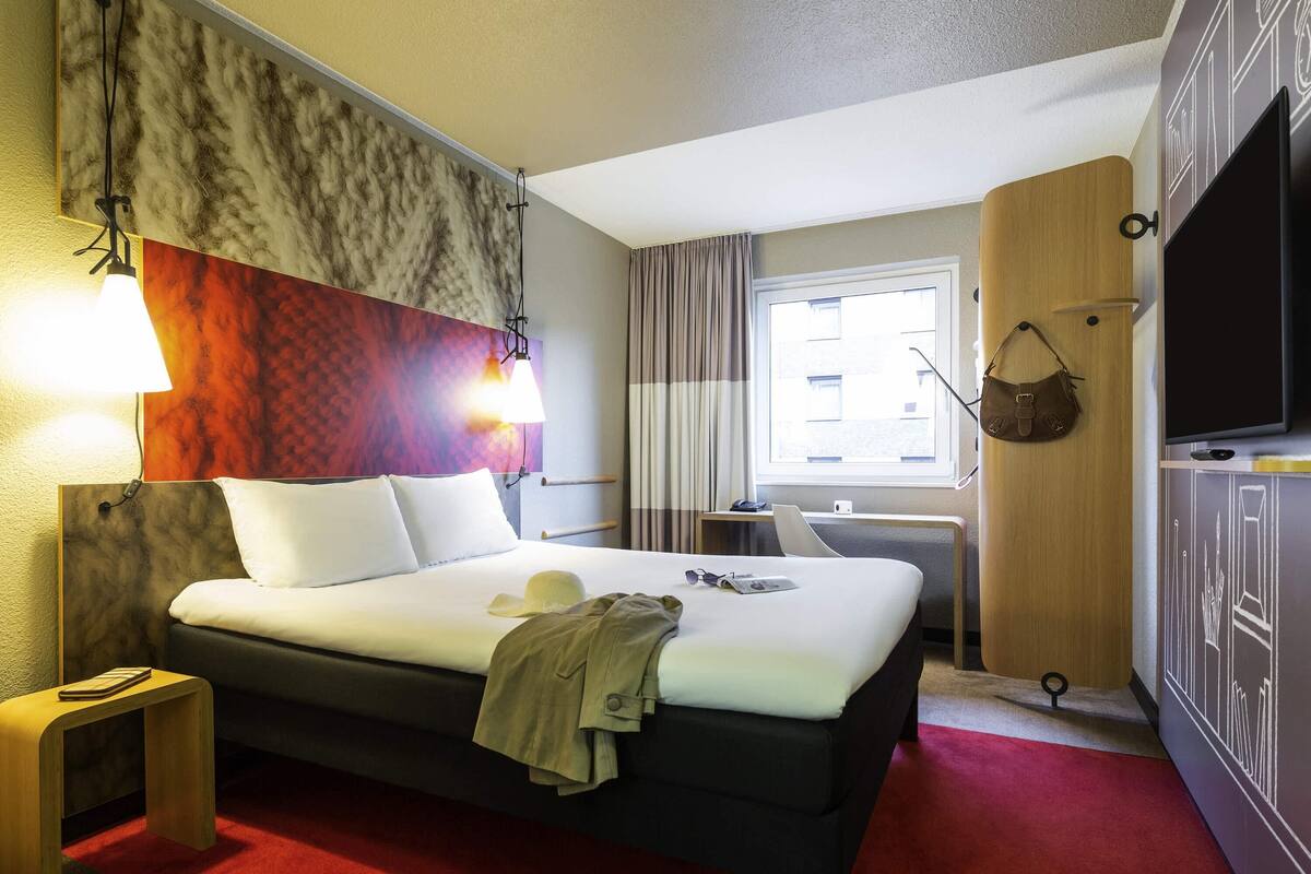 Une chambre d’hôtel comprenant un lit, des lampes de chevet, une télévision, une fenêtre avec des rideaux, un bureau et un porte-manteau.