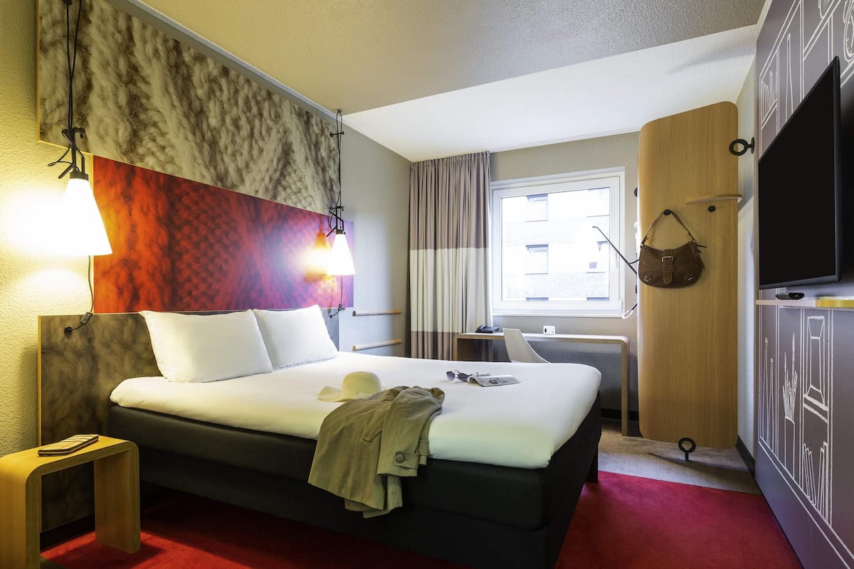 Habitación de hotel con cama, lámparas de noche, televisión, ventana con cortinas, escritorio y perchero.