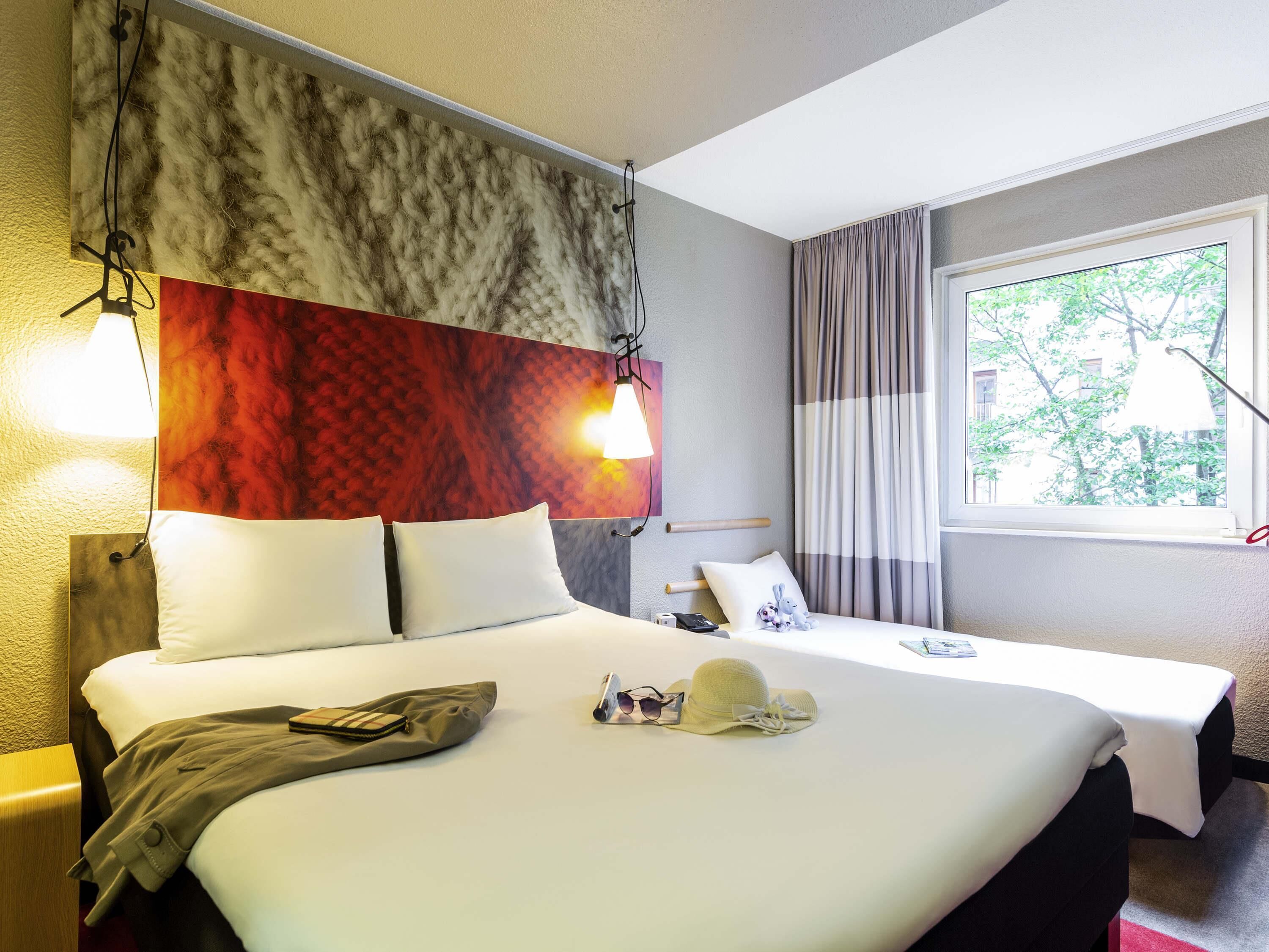 Une chambre d’hôtel avec un grand lit, une tête de lit au motif texturé, une fenêtre avec des rideaux et une vue sur les arbres à l’extérieur.