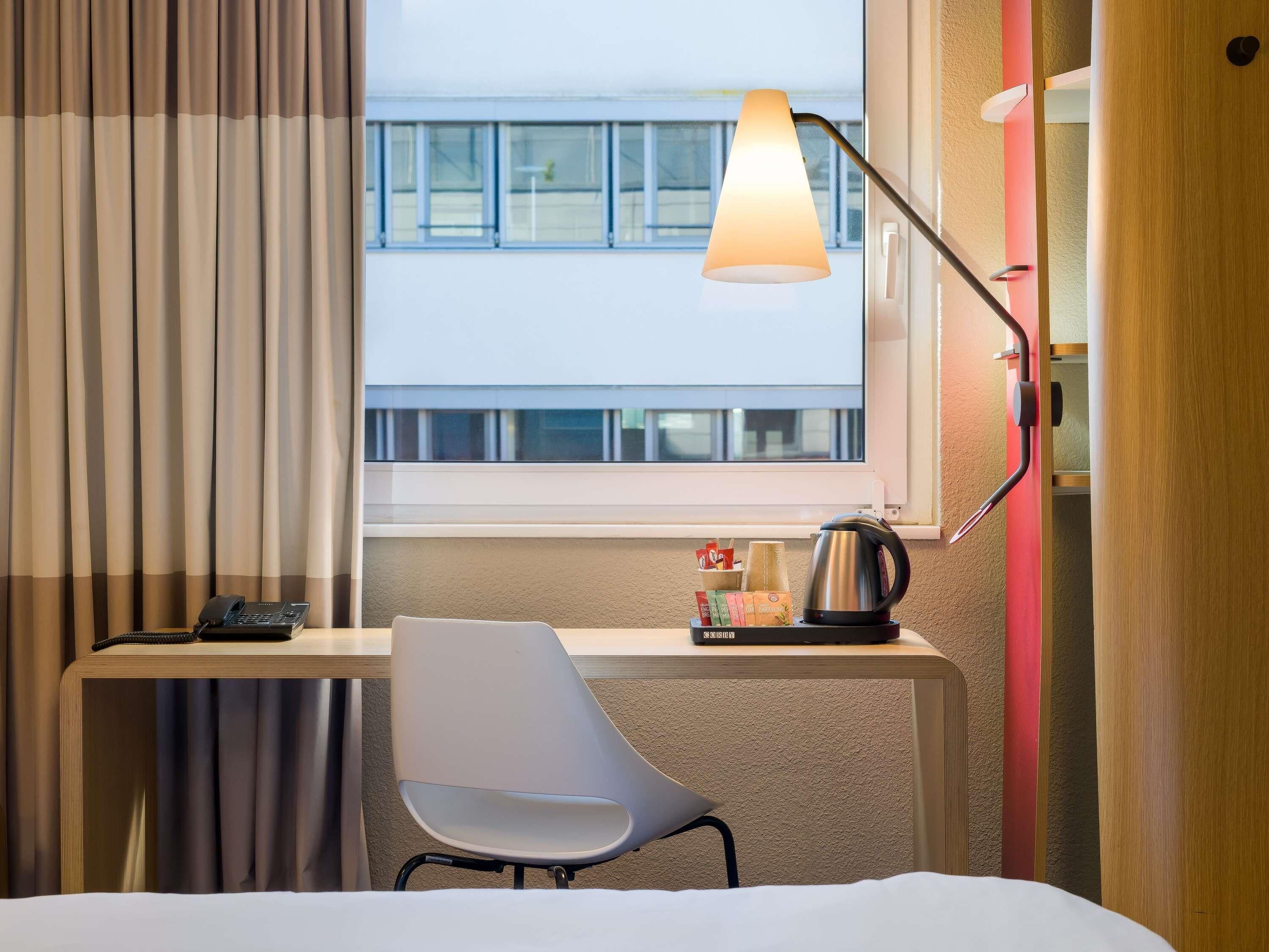A hotel room with a desk, chair, lamp, and a view of a building outside the window.