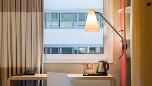 Une chambre d’hôtel équipée d’un bureau, d’une chaise, d’une lampe et offrant une vue sur un bâtiment situé à l’extérieur, visible par la fenêtre.