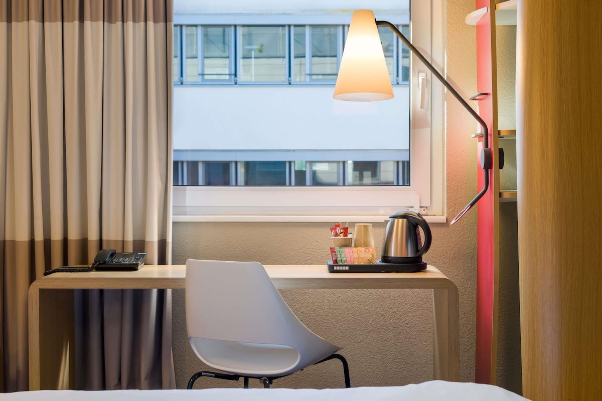 A hotel room with a desk, chair, lamp, and a view of a building outside the window.