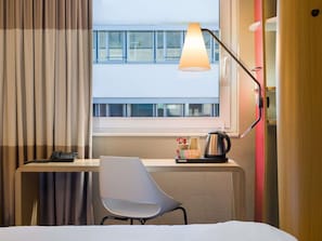 Une chambre d’hôtel équipée d’un bureau, d’une chaise, d’une lampe et offrant une vue sur un bâtiment situé à l’extérieur, visible par la fenêtre.