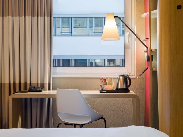 Une chambre d’hôtel équipée d’un bureau, d’une chaise, d’une lampe et offrant une vue sur un bâtiment situé à l’extérieur, visible par la fenêtre.