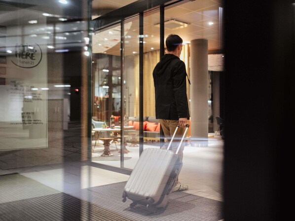 Un homme, une valise à la main, se trouve dans un hall d’entrée moderne aux murs de verre, avec un espace détente et une enseigne bien visible.