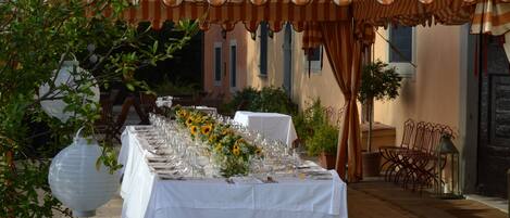 An outdoor dining area with a long table set for a meal, covered by a striped awning, surrounded by chairs and potted plants.