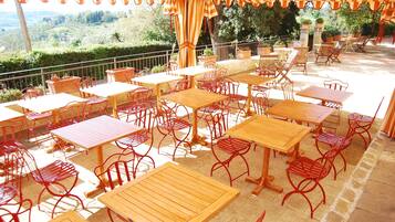 Un patio al aire libre con varias mesas de madera y sillas de metal rojo, rodeado de plantas en macetas y vistas a un paisaje pintoresco.