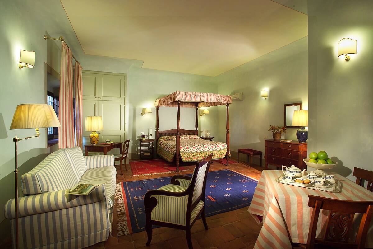 A bedroom with a four-poster bed, a sofa, a chair, a dining table with a tray of breakfast items, and a vase with flowers.