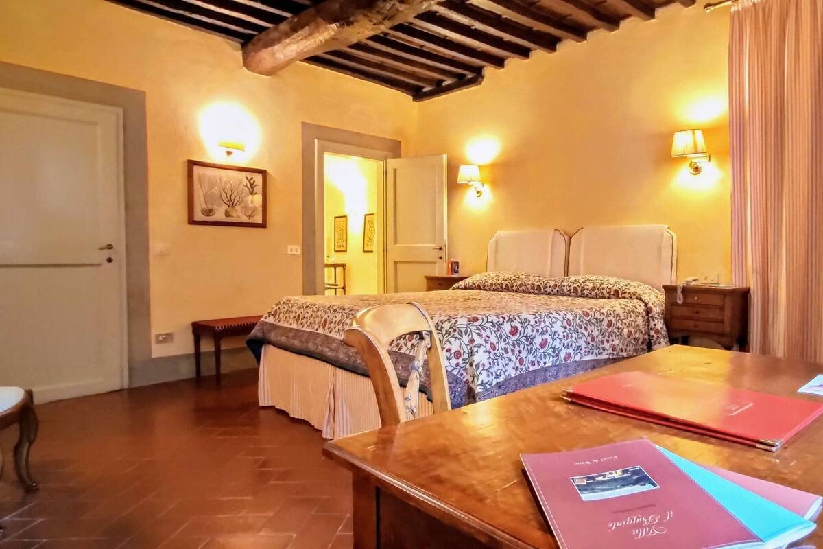 A bedroom with a bed, bedside tables, a wooden desk, and a chair. There are two bedside lamps, a door, and a wall-mounted picture.