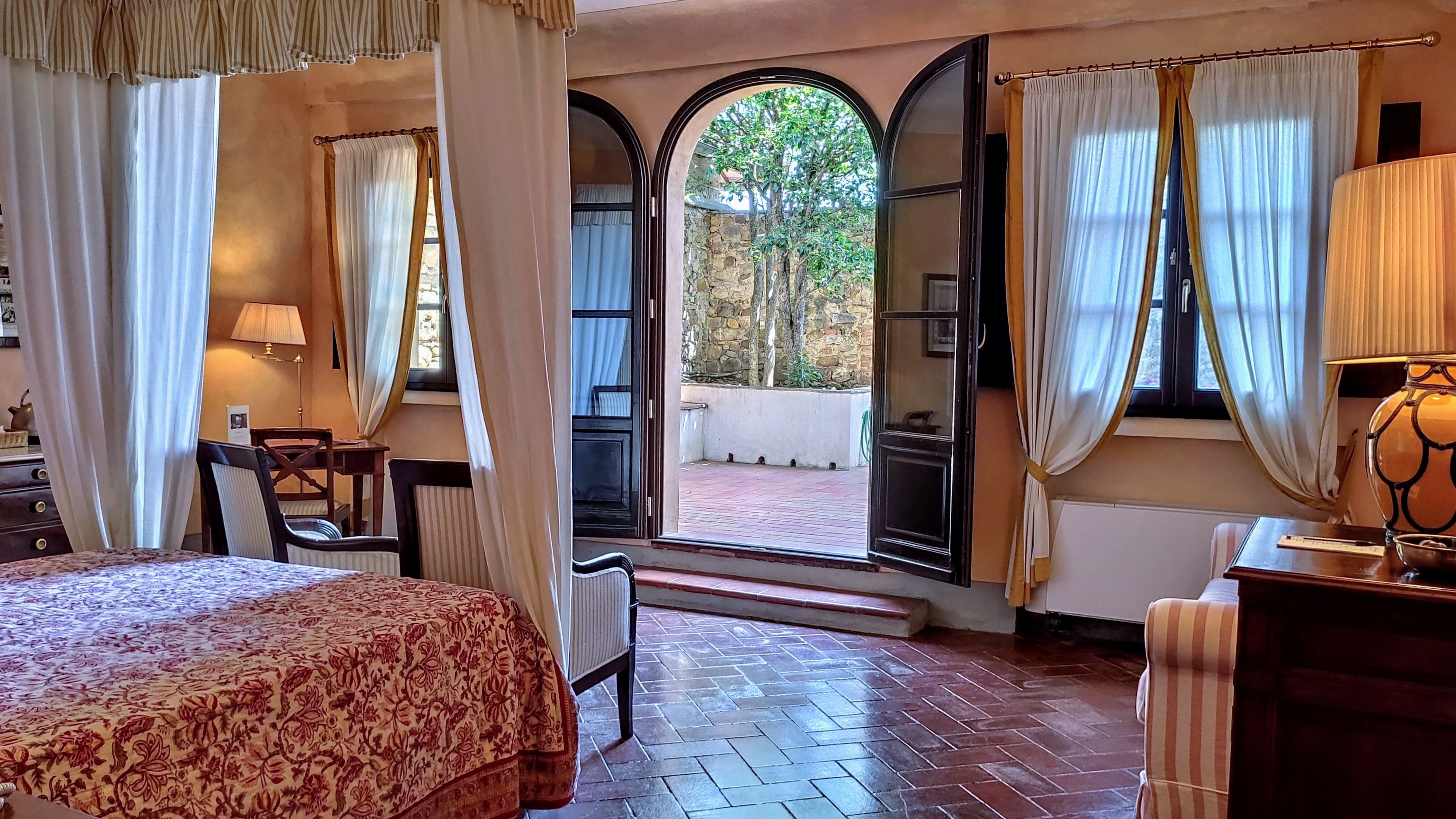 A hotel room with a bed, a desk, a chair, a lamp, and a view of a courtyard through an arched doorway.